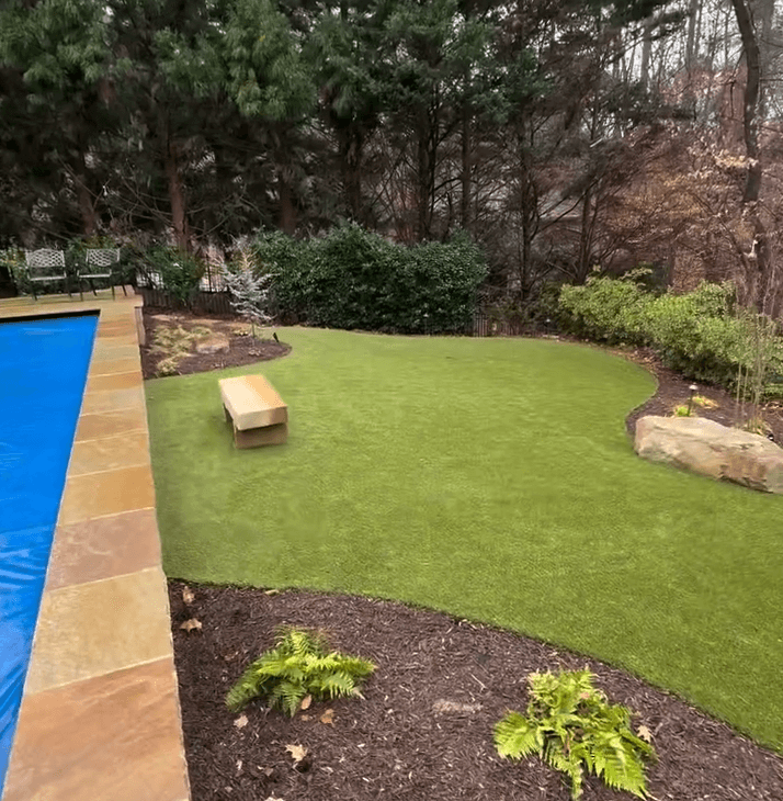 Backyard outdoor living space designed by a hardscaping and outdoor living contractor, featuring a pool, stone bench on artificial grass, landscaped mulch beds, and mature trees.