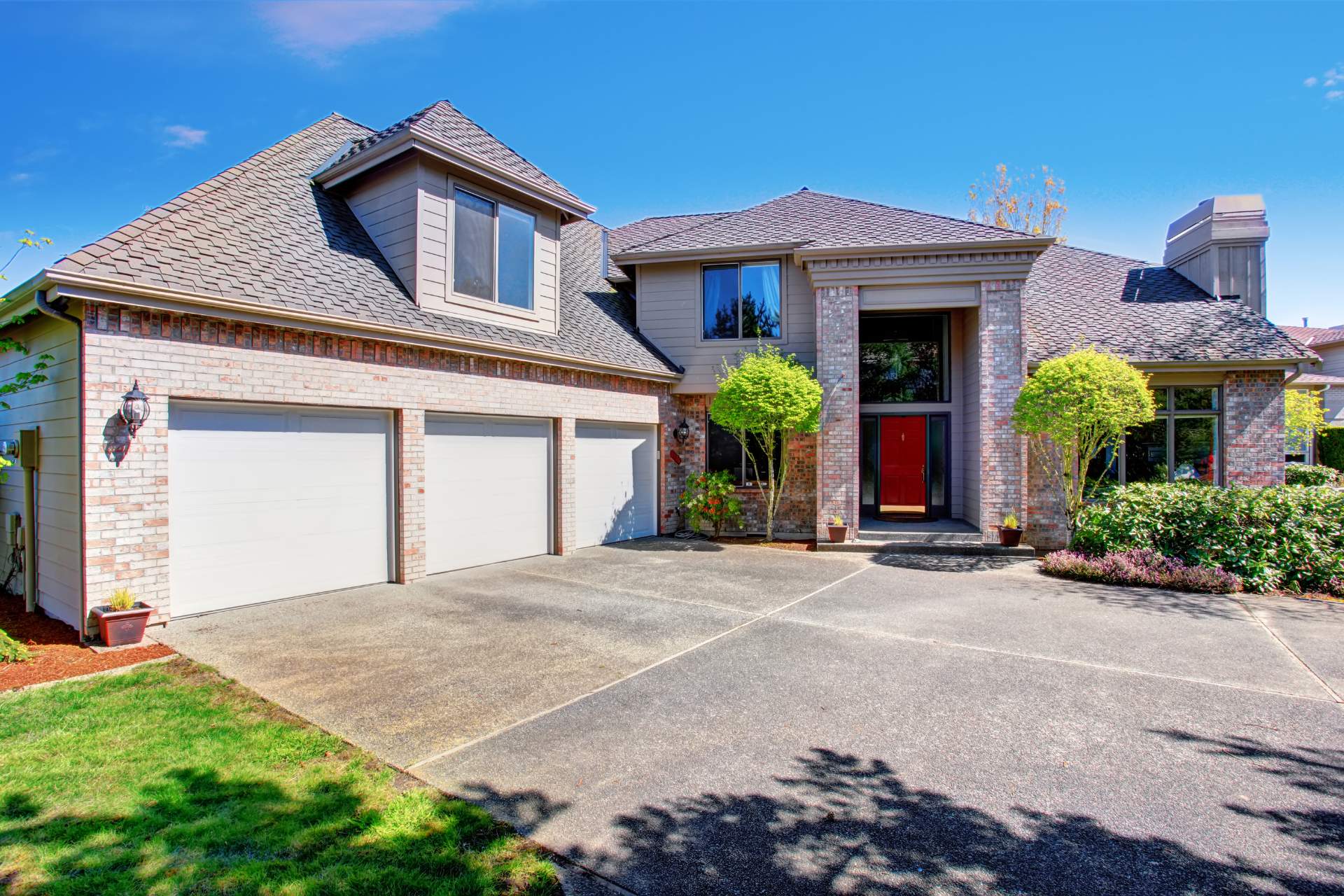 Two-story suburban home featuring concrete driveway installation, a brick-and-siding exterior with a three-car garage, red front door, large windows, landscaped front yard, and a custom fire pit installation under a clear blue sky.