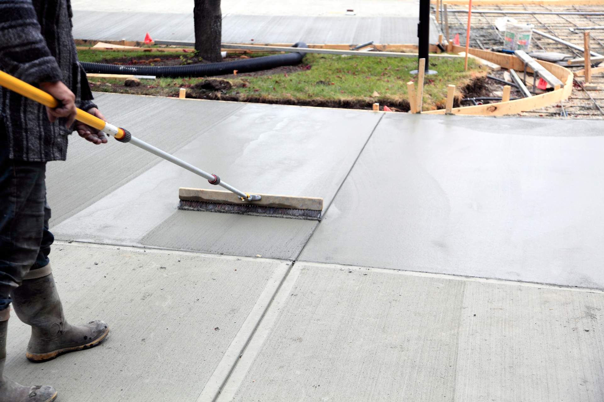 A worker uses a long-handled tool to smooth freshly poured concrete on a sidewalk under construction, preparing the surface for possible features like an outdoor fireplace installation.