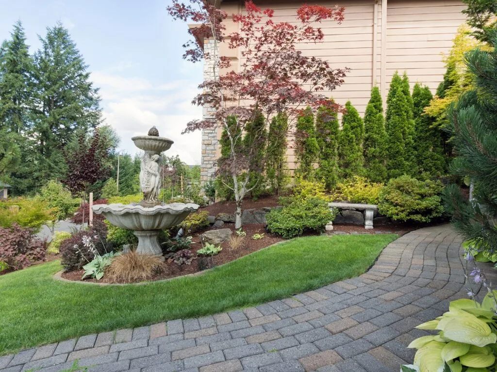 A neatly landscaped garden featuring hardscaping ideas for backyard spaces, with a stone fountain, curved walkway, manicured lawn, various shrubs, and a wooden bench beside the house.