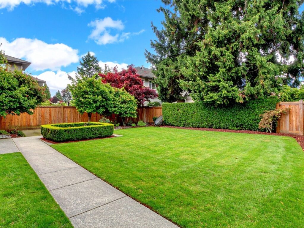 A fenced backyard features a neatly mowed lawn, trimmed hedges, several trees, and a concrete walkway—perfect inspiration if you’re looking for hardscaping ideas for backyard spaces under a partly cloudy sky.
