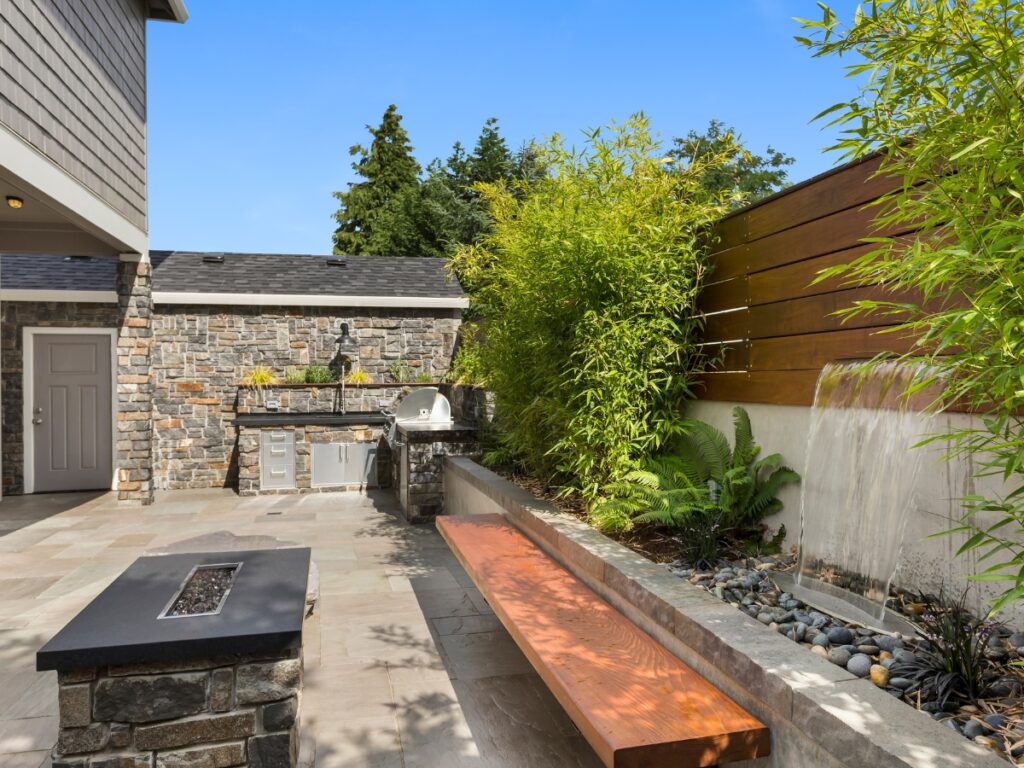 Modern backyard patio showcasing hardscaping ideas for backyard: stone outdoor kitchen, grill, wooden bench, fire pit, bamboo plants, and a built-in wall fountain, all bathed in sunlight.