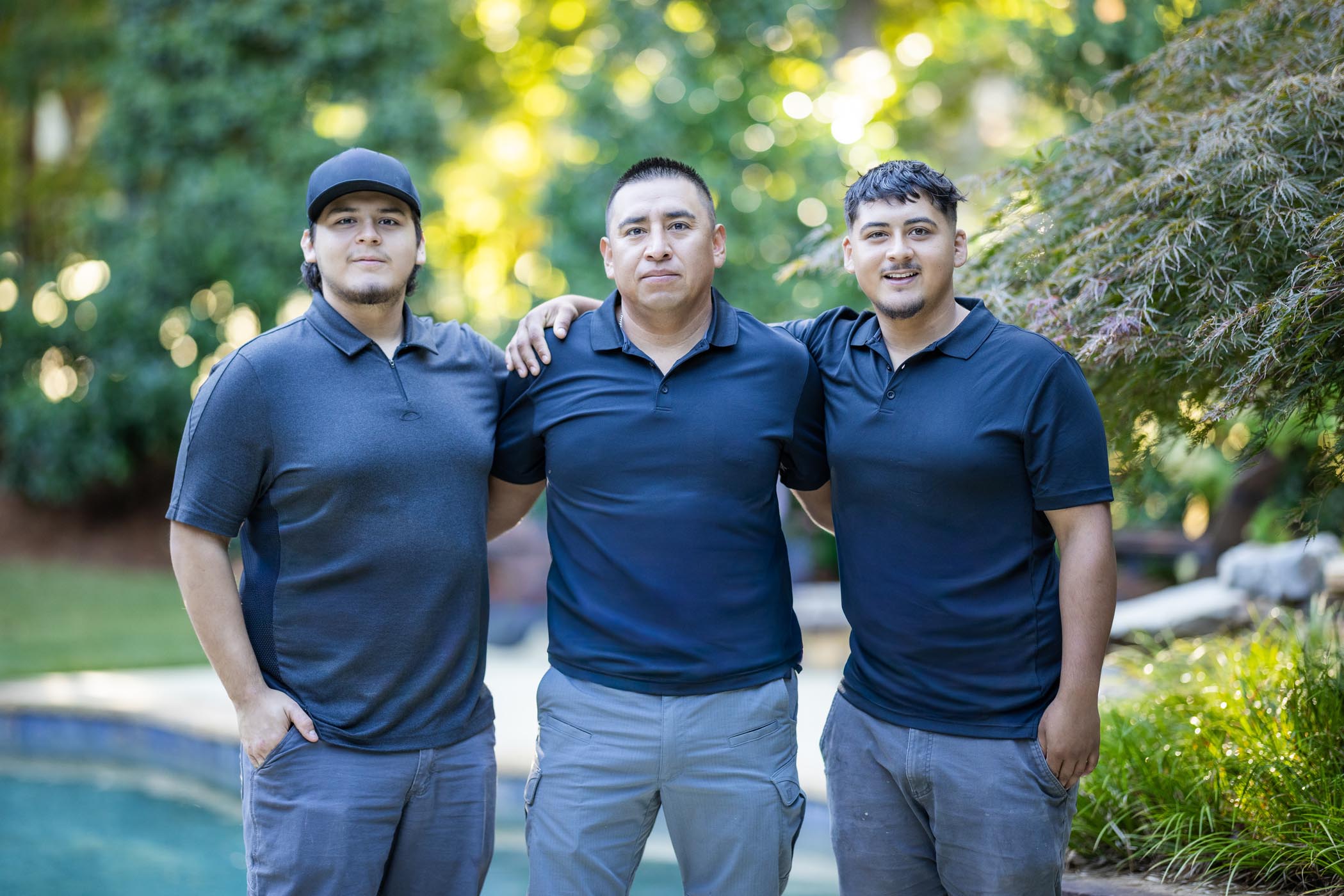 Three men wearing navy polo shirts and gray pants stand outdoors together, with two of them resting their hands on the shoulders of the man in the center. Greenery is visible in the background.