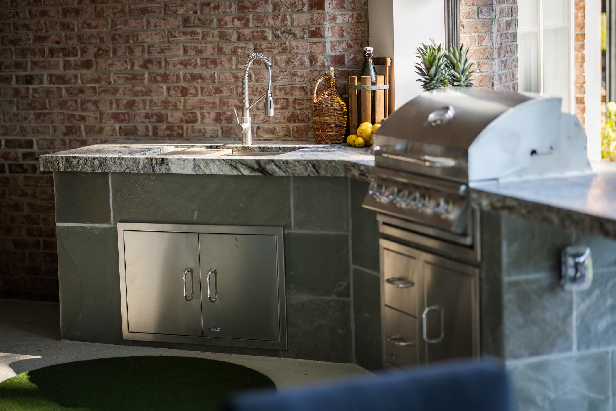Outdoor kitchen with a stainless steel grill, sink with a gooseneck faucet, marble countertop, and decorative items including a pineapple and a basket of lemons.