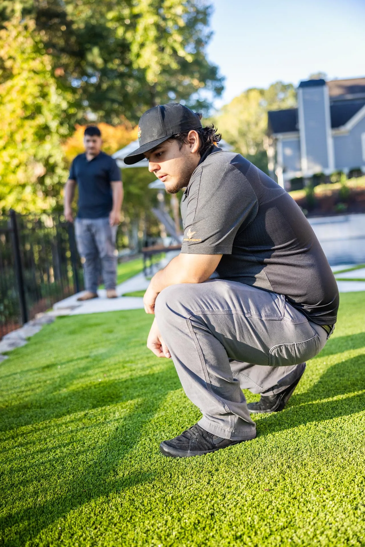 A man in a black cap and gray pants squats on artificial grass while another man stands in the background near a fence and trees.