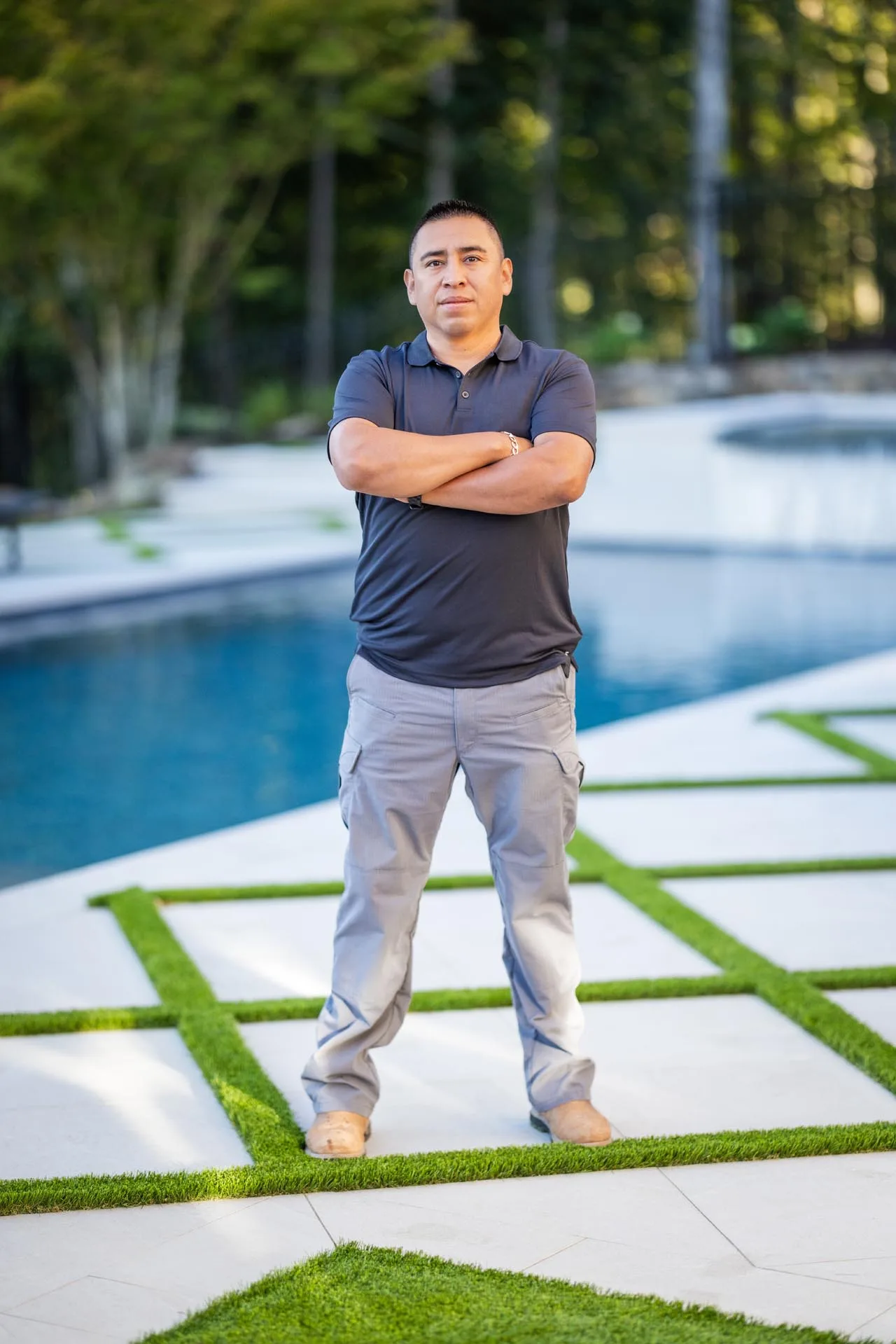 A man stands with arms crossed on a geometric patio near a pool, wearing a dark polo shirt, gray pants, and tan shoes. Trees and greenery are visible in the background.