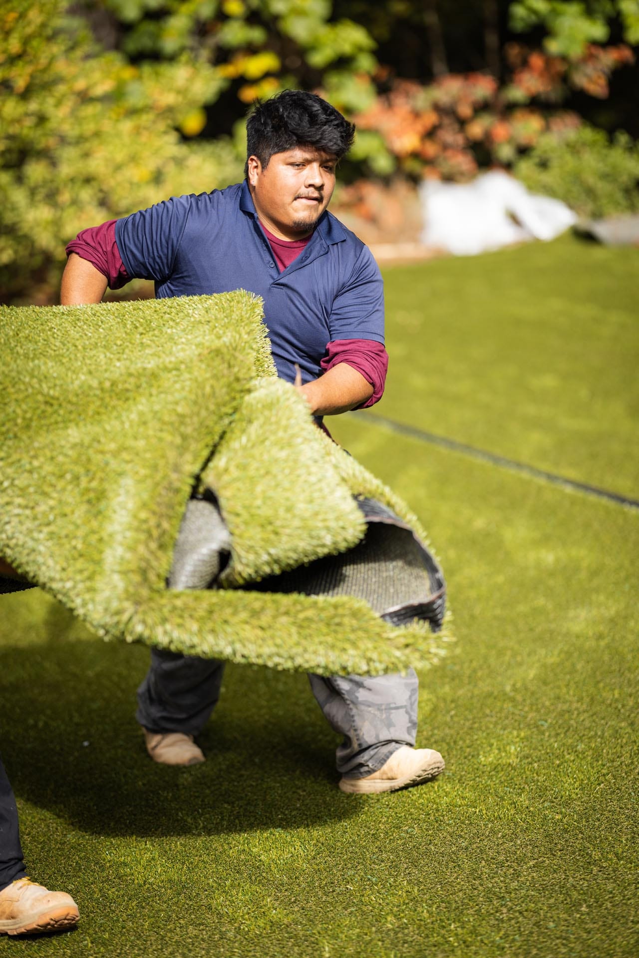 A person in work clothes lifts and moves a large roll of artificial grass on a lawn, with trees and plants in the background.
