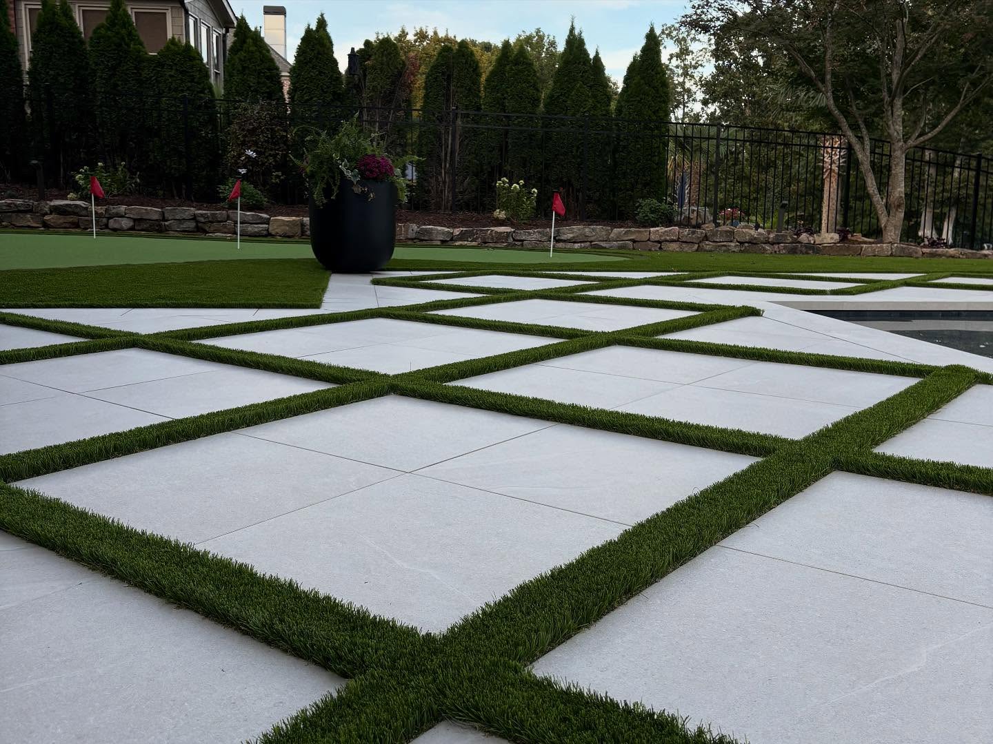 A backyard with large square white pavers arranged in a grid pattern, separated by strips of grass, next to a putting green with small red flags and landscaping in the background.