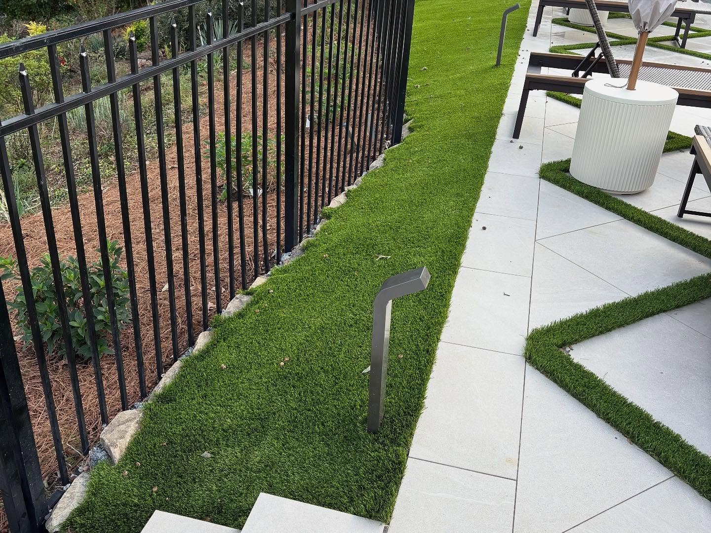 A black metal fence borders a landscaped area with mulch and plants; artificial grass and modern lighting run alongside tiled patio flooring with lounge chairs.
