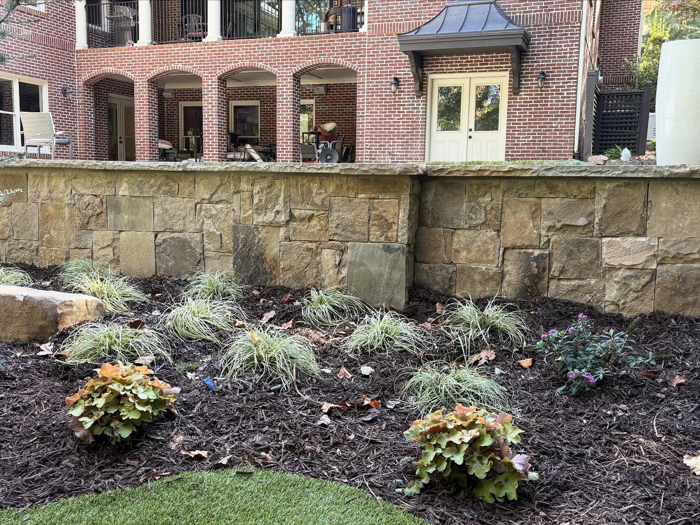 A landscaped garden bed with small shrubs and grasses mulched in front of a stone retaining wall; brick house with patio and balconies in the background.