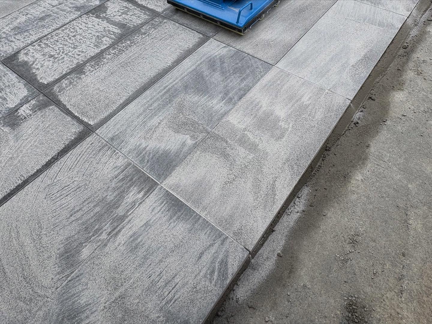 Gray paving stones partially covered with fine sand are arranged beside a concrete surface. A blue plastic object sits on top of the pavers in the upper part of the image.