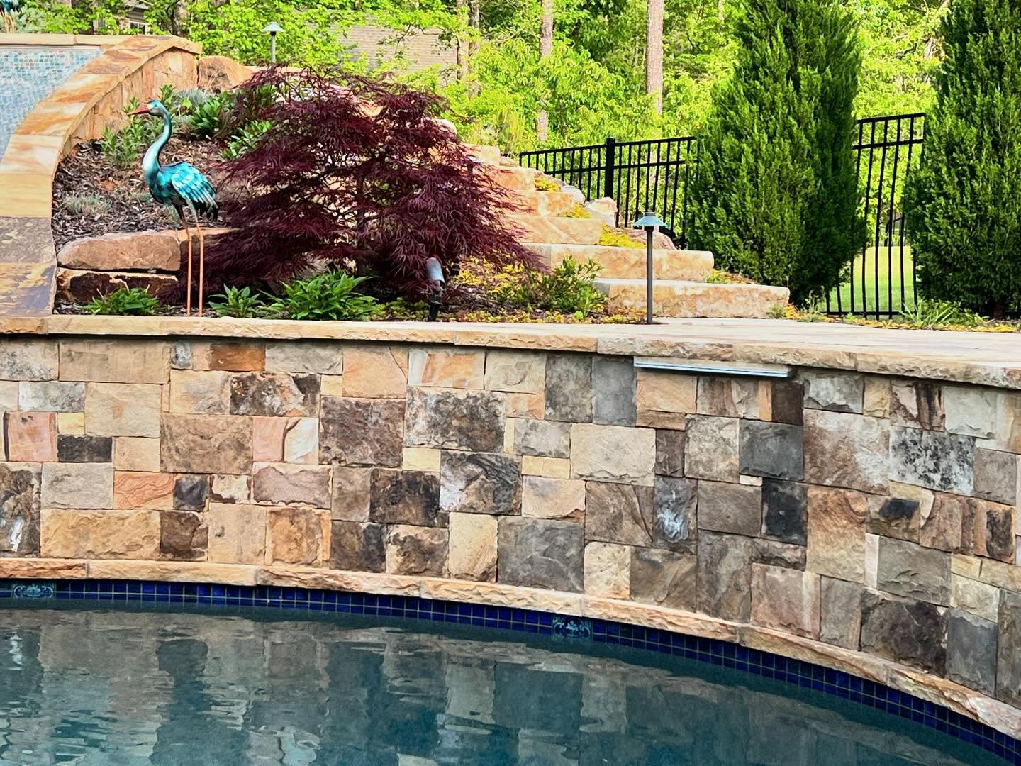 A stone retaining wall borders a pool, with landscaped plants, a metal bird sculpture, and stone steps leading up to a grassy area and black metal fence.