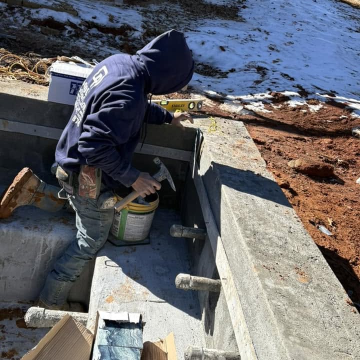 A person in a hoodie uses a level and hammer while working on a concrete structure at a construction site with snow and exposed soil nearby.