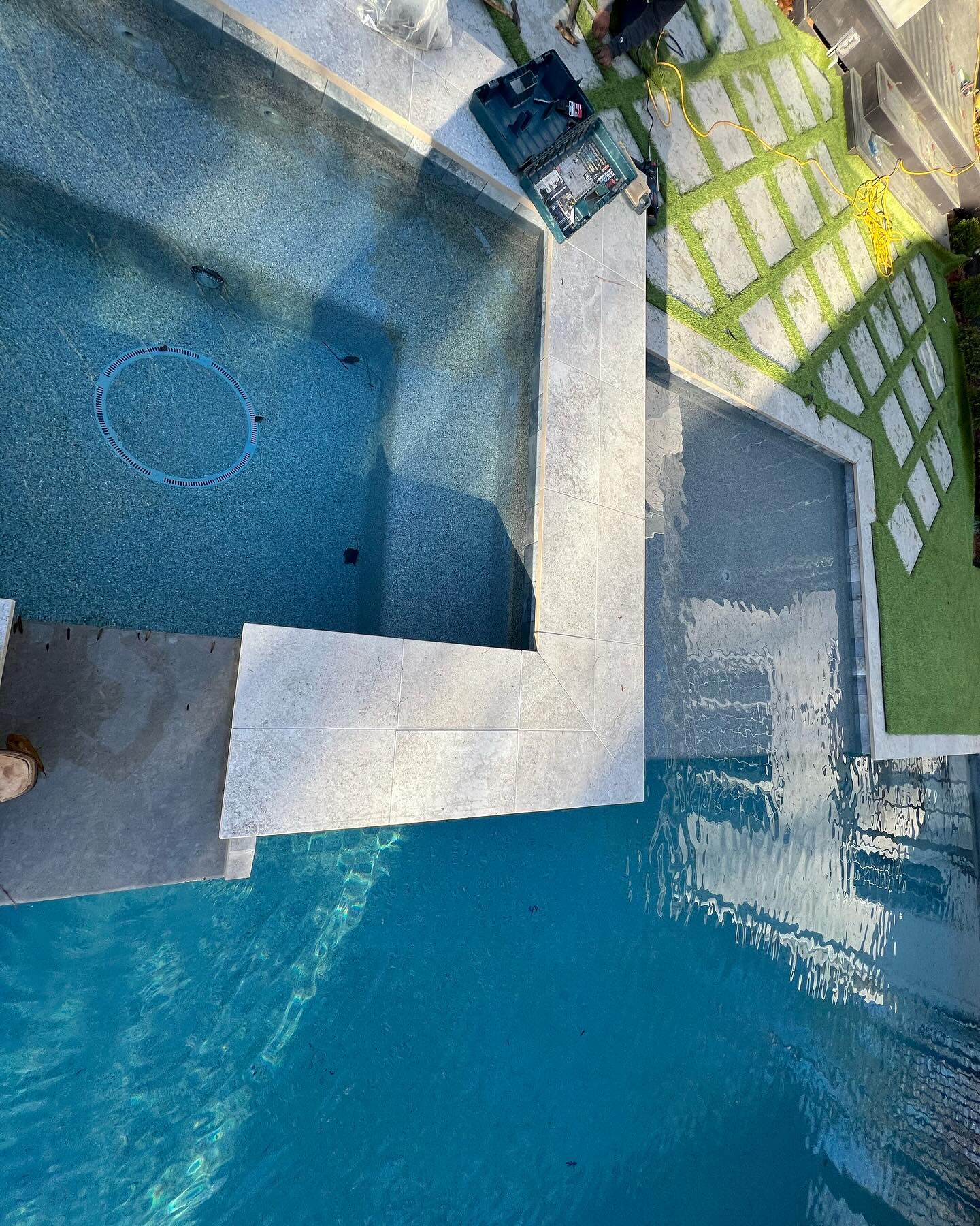 A swimming pool with a built-in hot tub, stone tile decking, and a grassy area with concrete pavers beside the pool. Tools and equipment are visible near the edge.