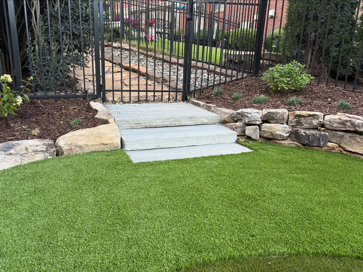 Stone steps lead from artificial grass to a fenced gate, bordered by a stone retaining wall and landscaped garden beds.