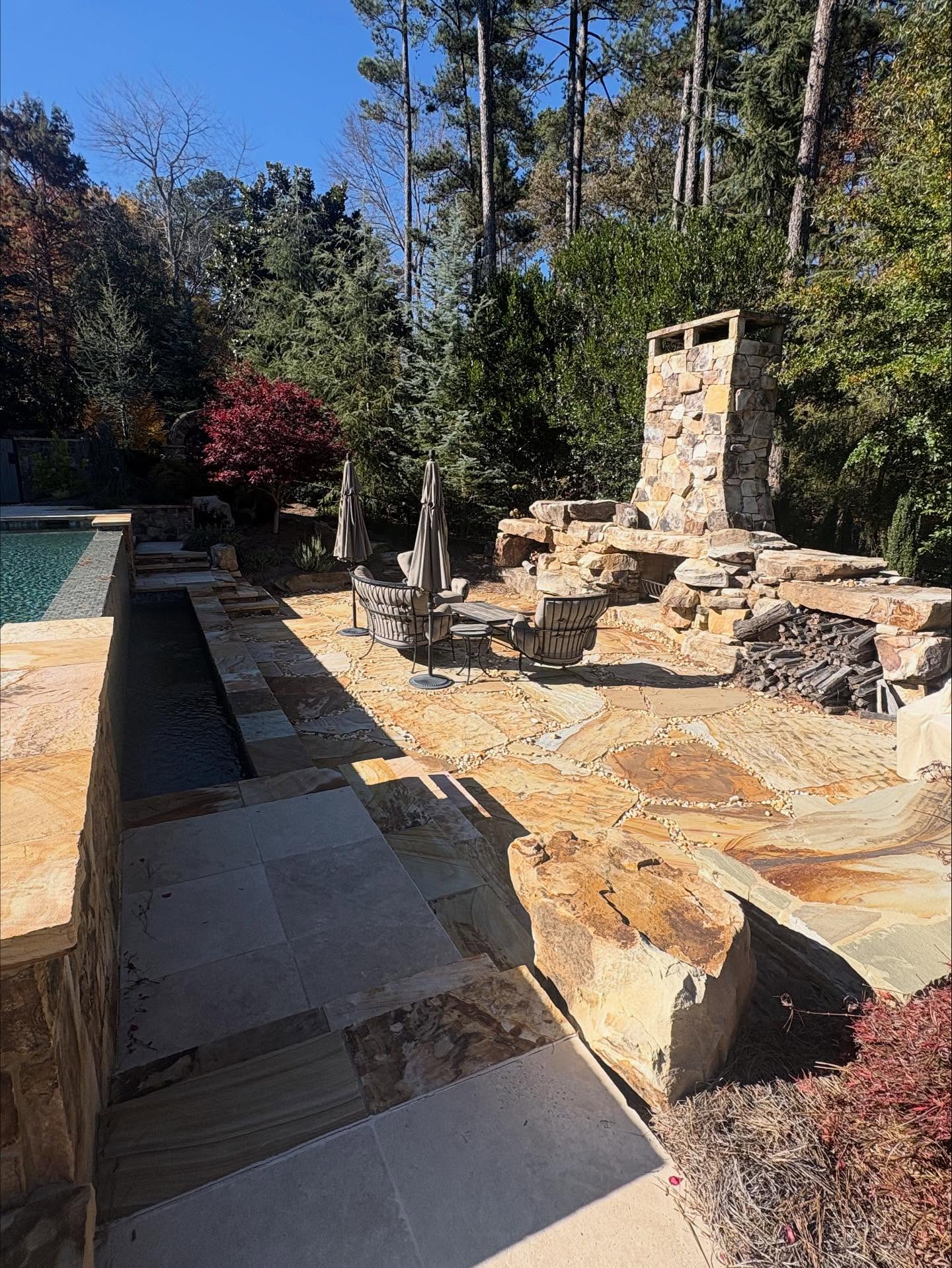 Stone patio area with round table, chairs, umbrellas, and an outdoor fireplace next to a pool, surrounded by trees and landscaping on a clear day.