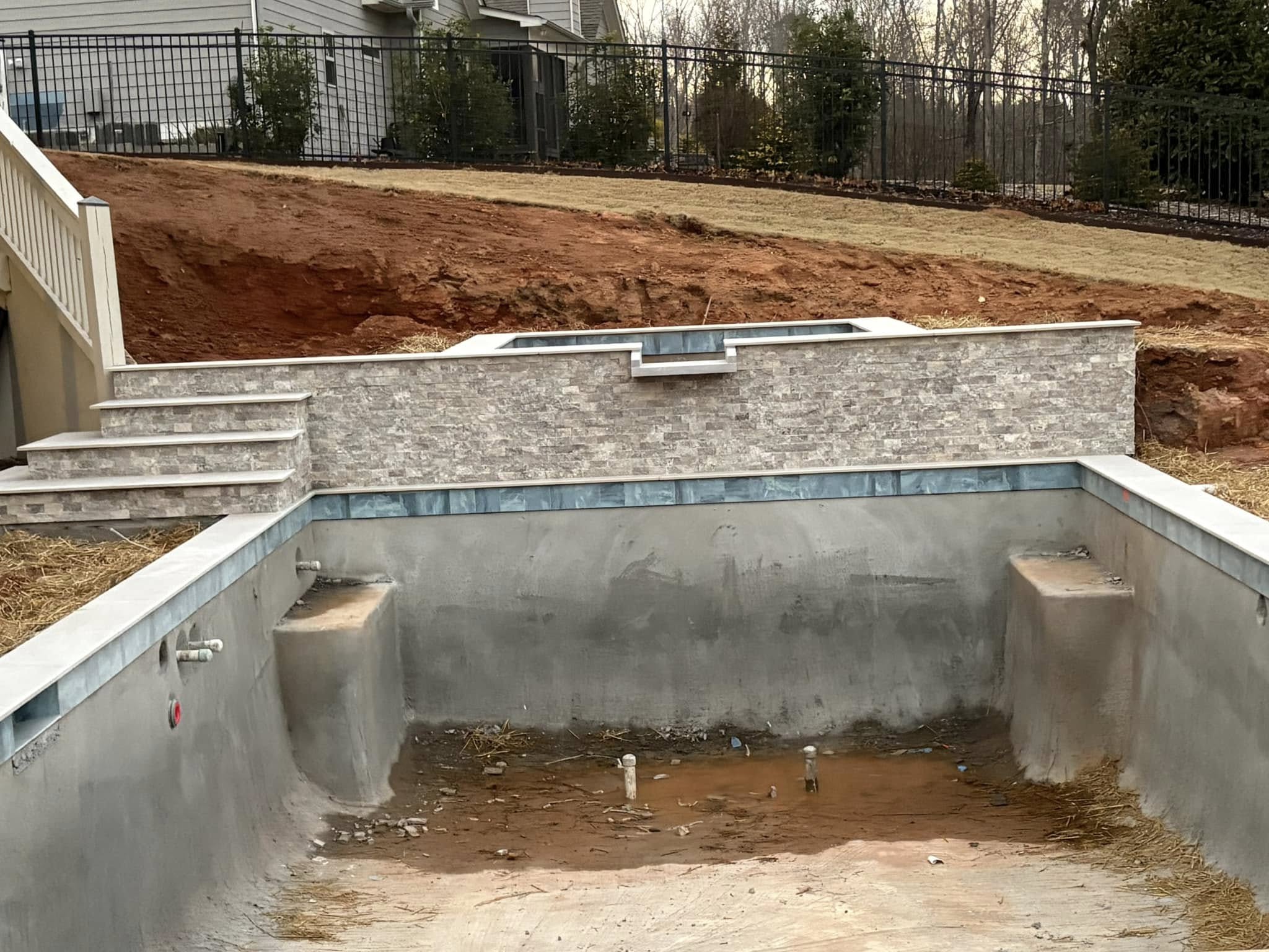 Unfinished swimming pool with exposed concrete and dirt, set below a retaining wall and stairs, in a backyard under construction.