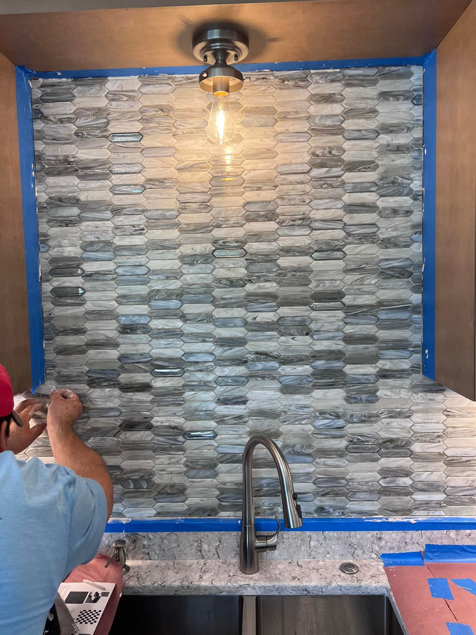 A person installs gray and white glass tile backsplash above a kitchen sink, with painter’s tape outlining the area and a light fixture overhead.