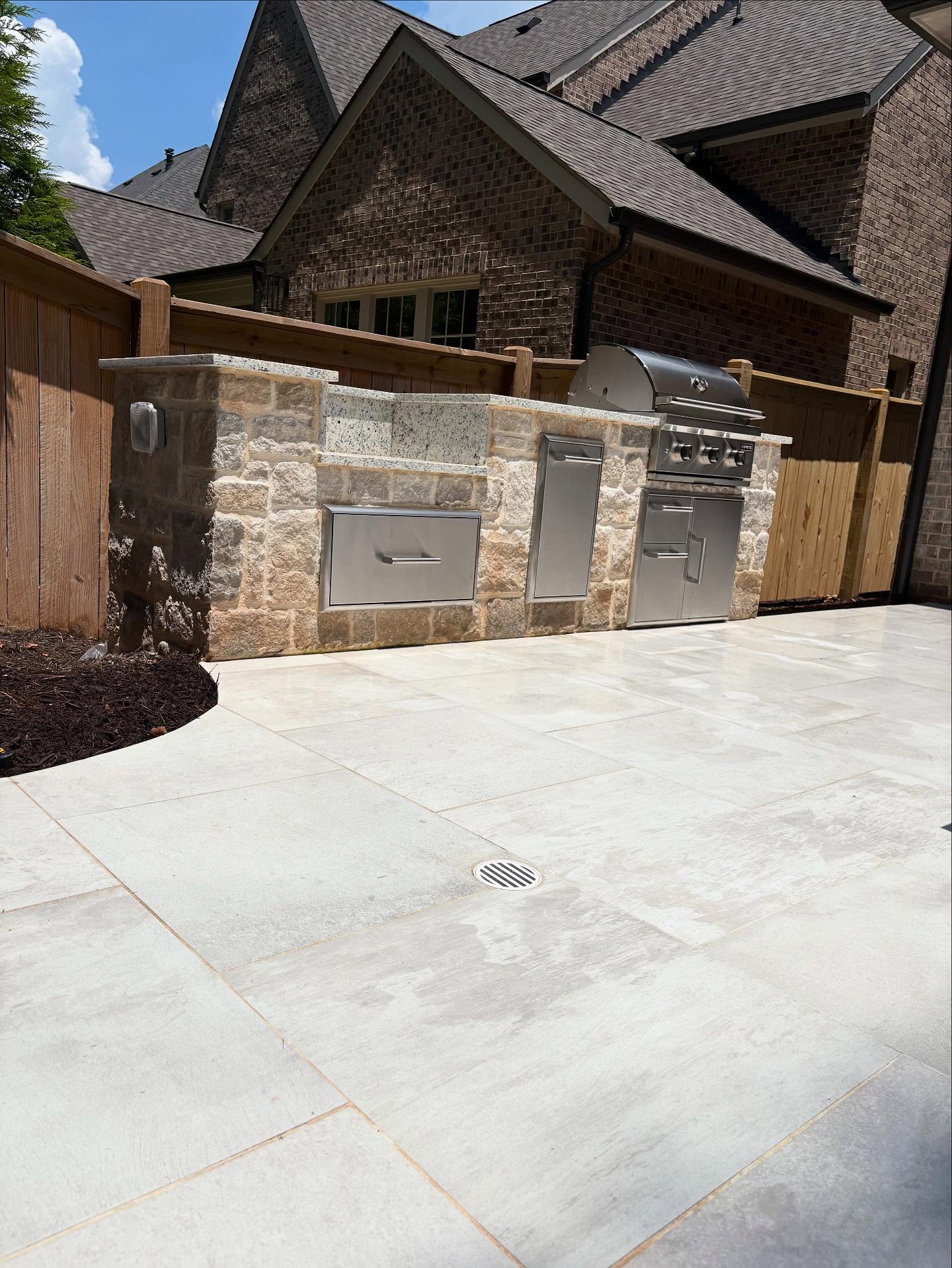 Outdoor stone kitchen with stainless steel grill, storage drawers, and cabinets, set against a wooden fence and brick house on a tiled patio.