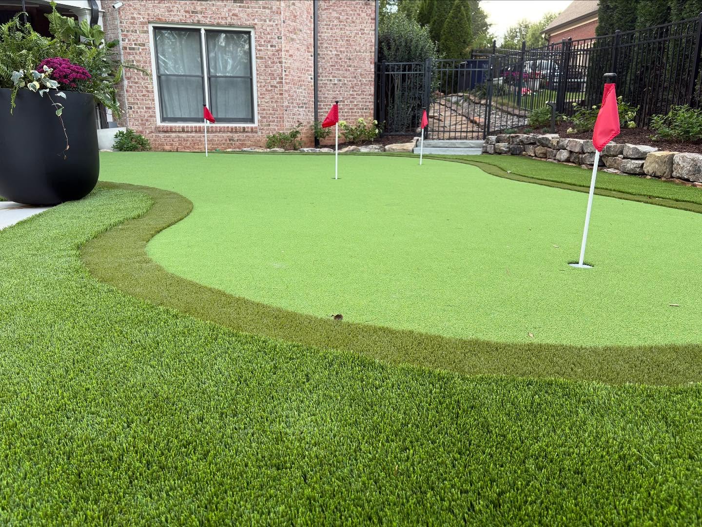 Artificial turf putting green with red flags, bordered by manicured grass and modern landscaping elements, enhancing outdoor living space for entertainment.