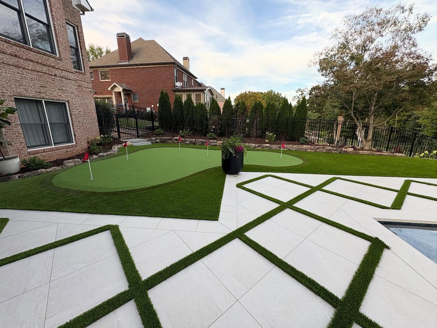 A backyard with a putting green, artificial grass, geometric white patio tiles, and a pool, bordered by landscaped plants and neighboring houses.
