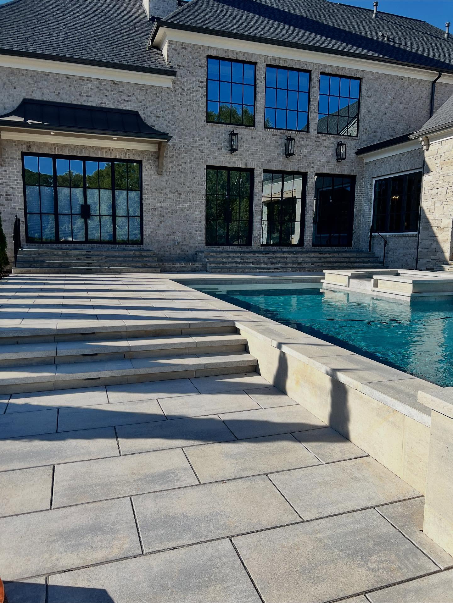 Large modern house with light brick exterior, large windows, and a swimming pool with wide stone steps leading up to the patio area.