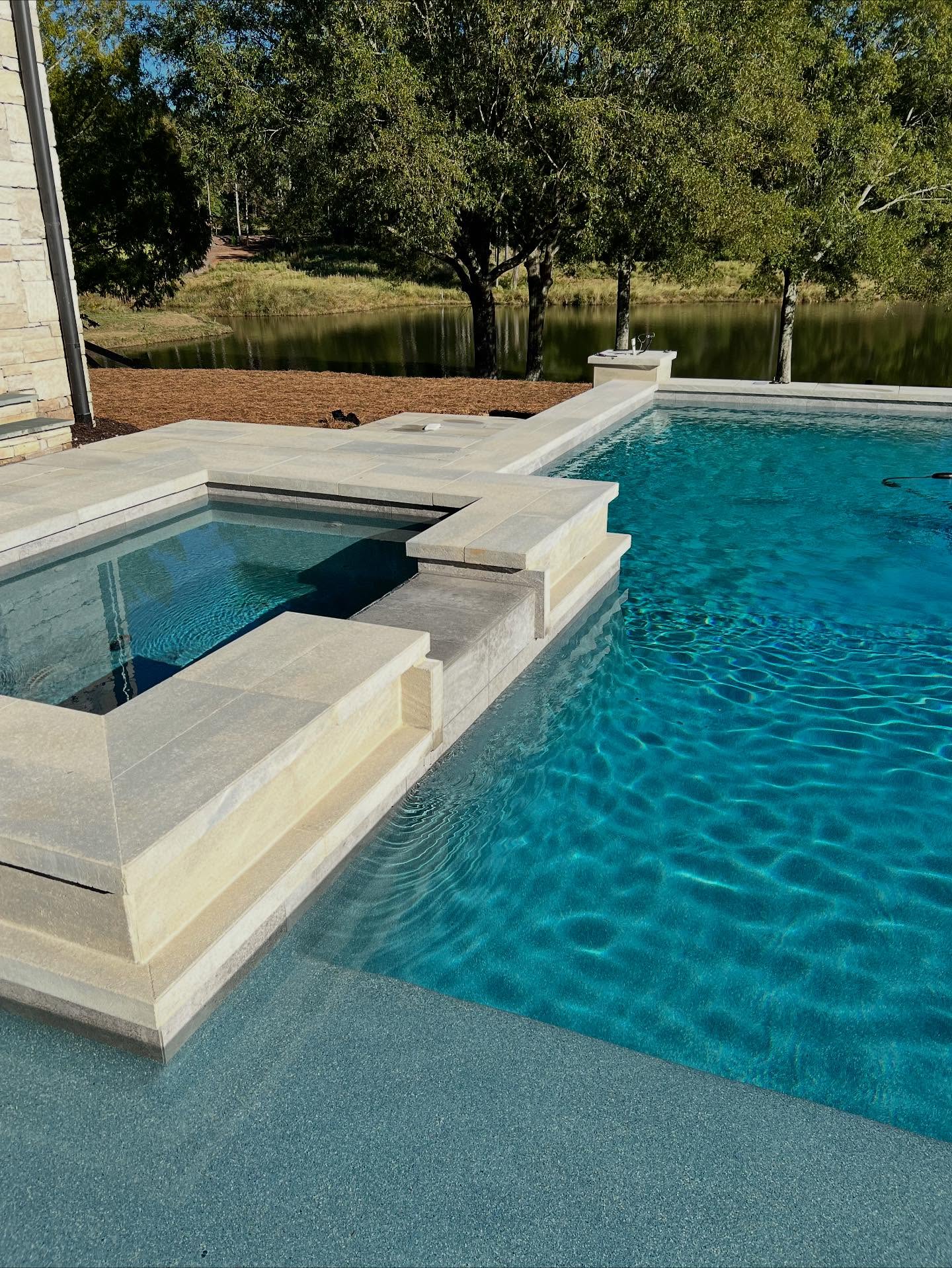A modern outdoor swimming pool with an attached square hot tub, stone steps, and clear blue water, set beside a pond and surrounded by trees.