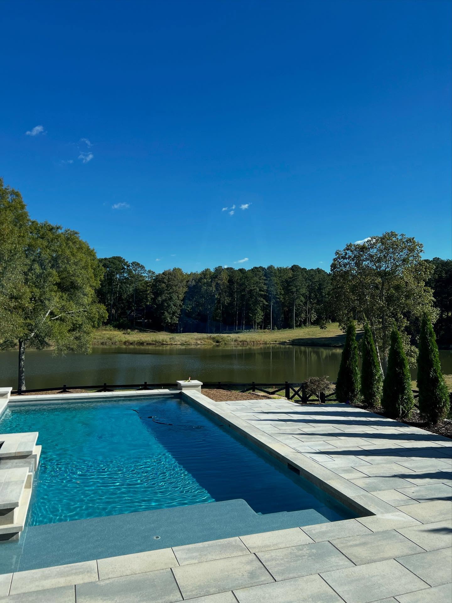 Rectangular outdoor pool with stone deck overlooking a calm lake, surrounded by trees under a clear blue sky.