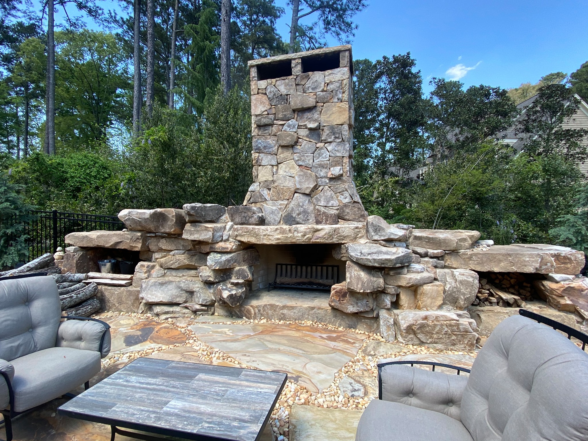Outdoor stone fireplace with a chimney surrounded by cushioned patio furniture and trees in the background.