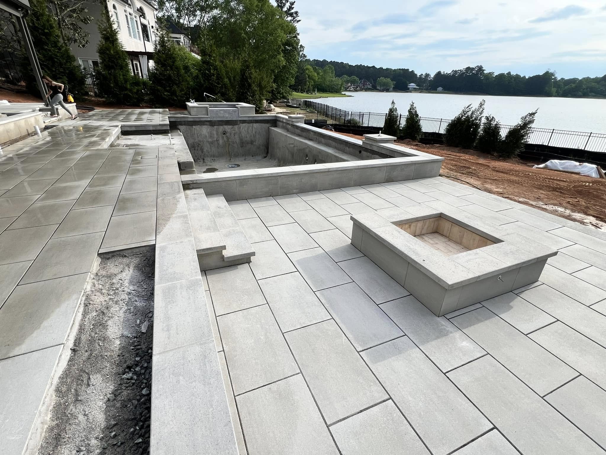 A newly constructed patio with large light gray pavers, an empty pool, a built-in fire pit, and a view of a lake in the background.