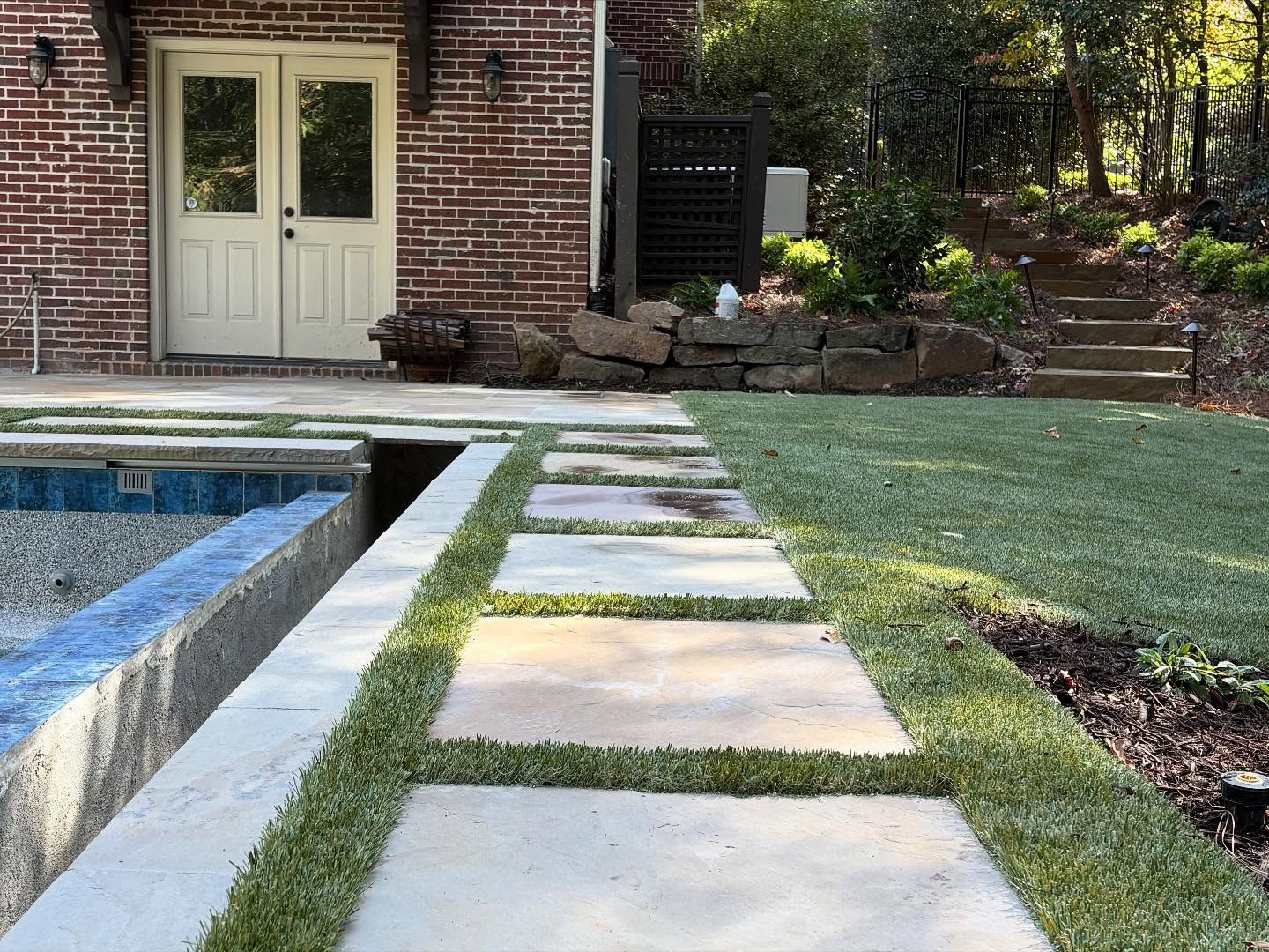 Stone pavers set in grass lead past an empty pool to a brick house with double doors; stone steps and landscaping appear on the right.