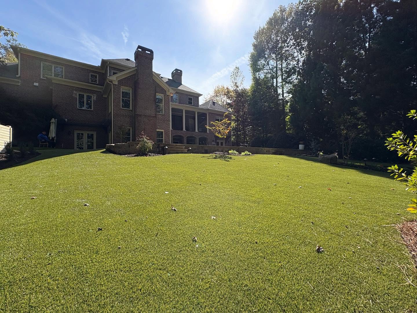 A large brick house with multiple chimneys overlooks a well-maintained grassy lawn, surrounded by trees under a bright sun.