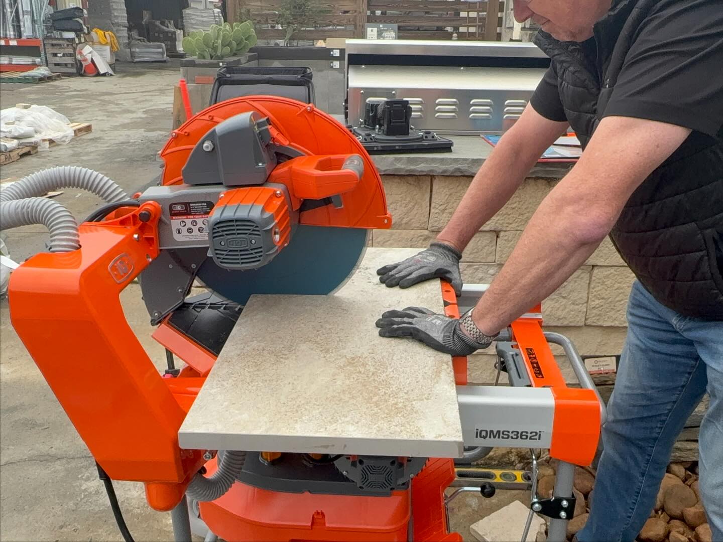 A person uses an orange iQMS362i saw to cut a large tile on a construction site, wearing gloves and standing next to a stone wall.