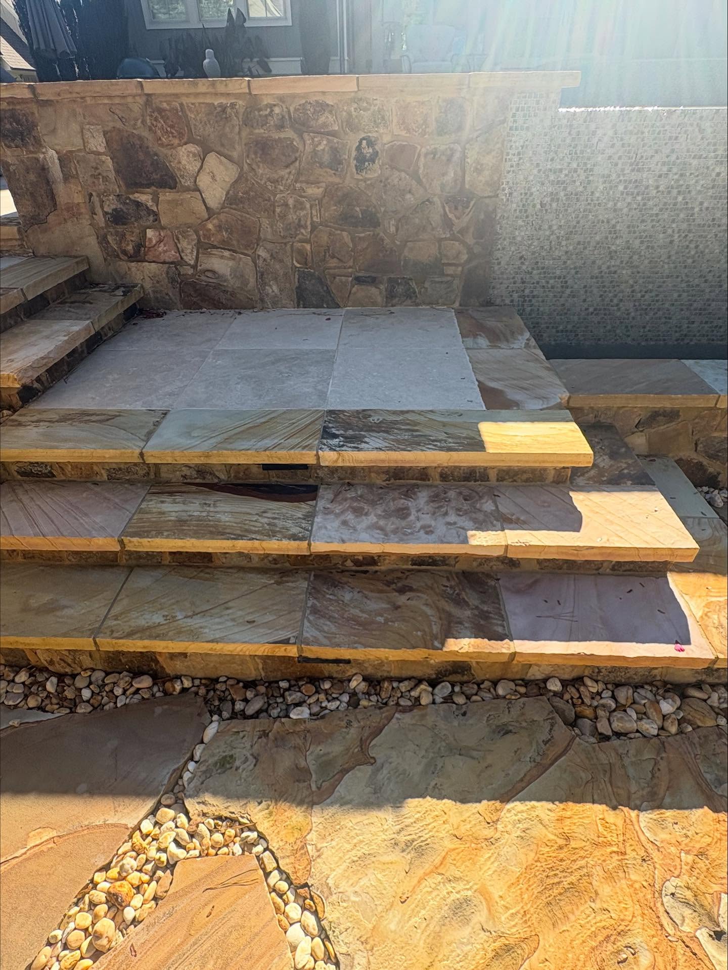 Three stone steps lead to a patio area with large stone tiles and a stone wall, partially in sunlight. Pebbles fill the gaps around the steps and paving.