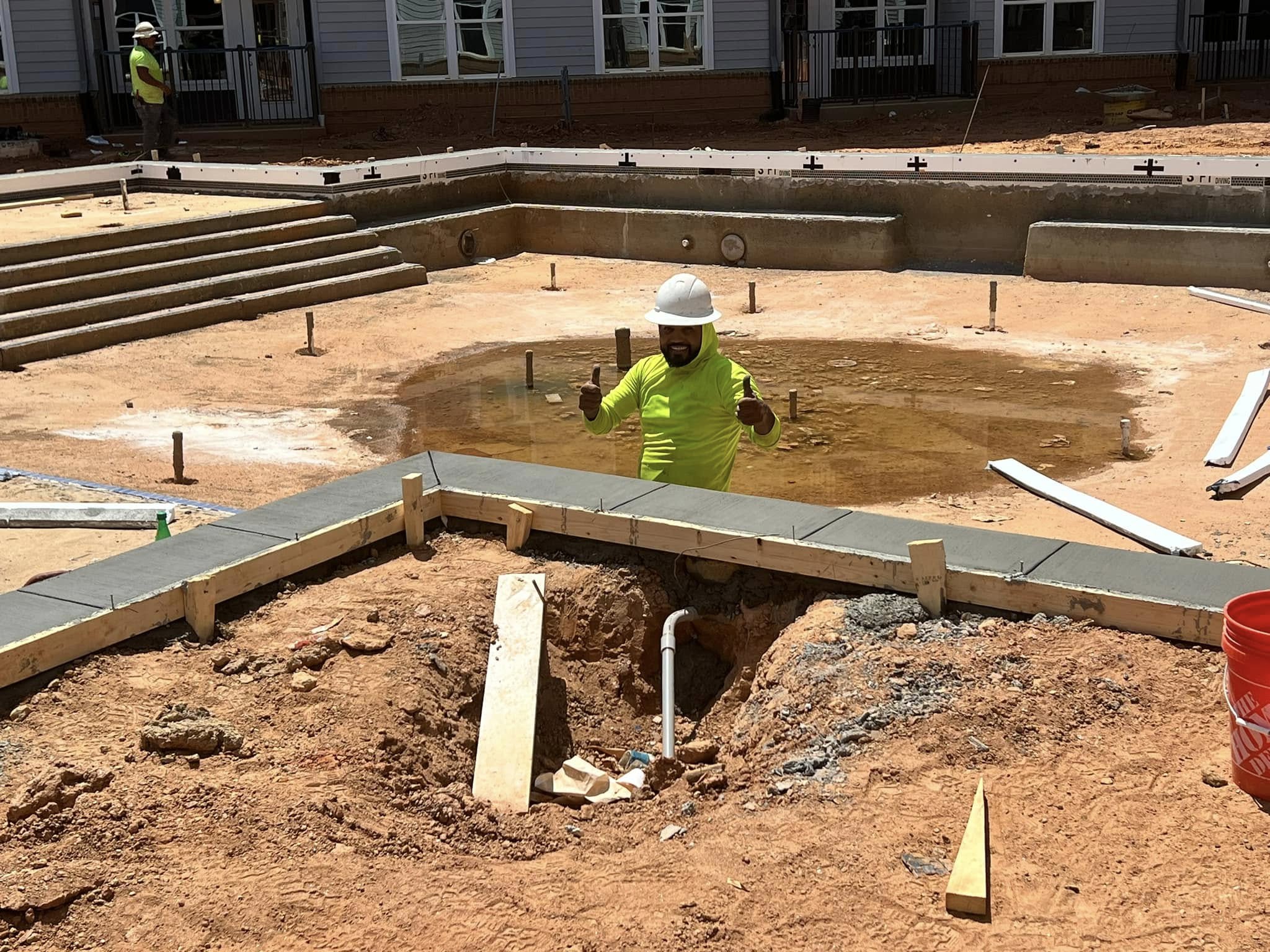 A construction worker in a neon shirt and hard hat stands in a muddy pit near a partially built pool surrounded by dirt and construction materials.