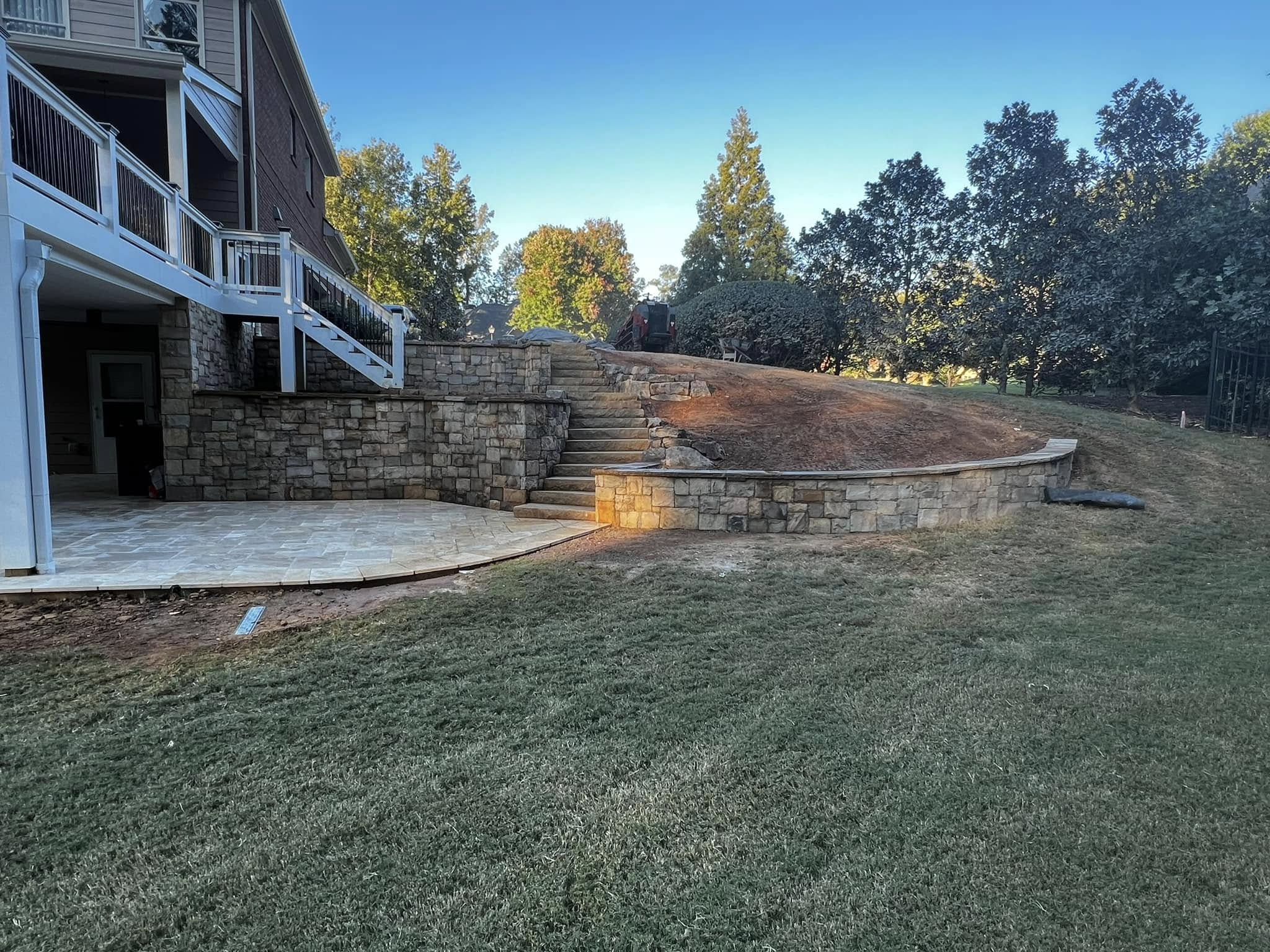 Stone retaining walls and steps create a tiered landscape behind a house, with unfinished earth on the upper level and a paved patio area on the lower level.