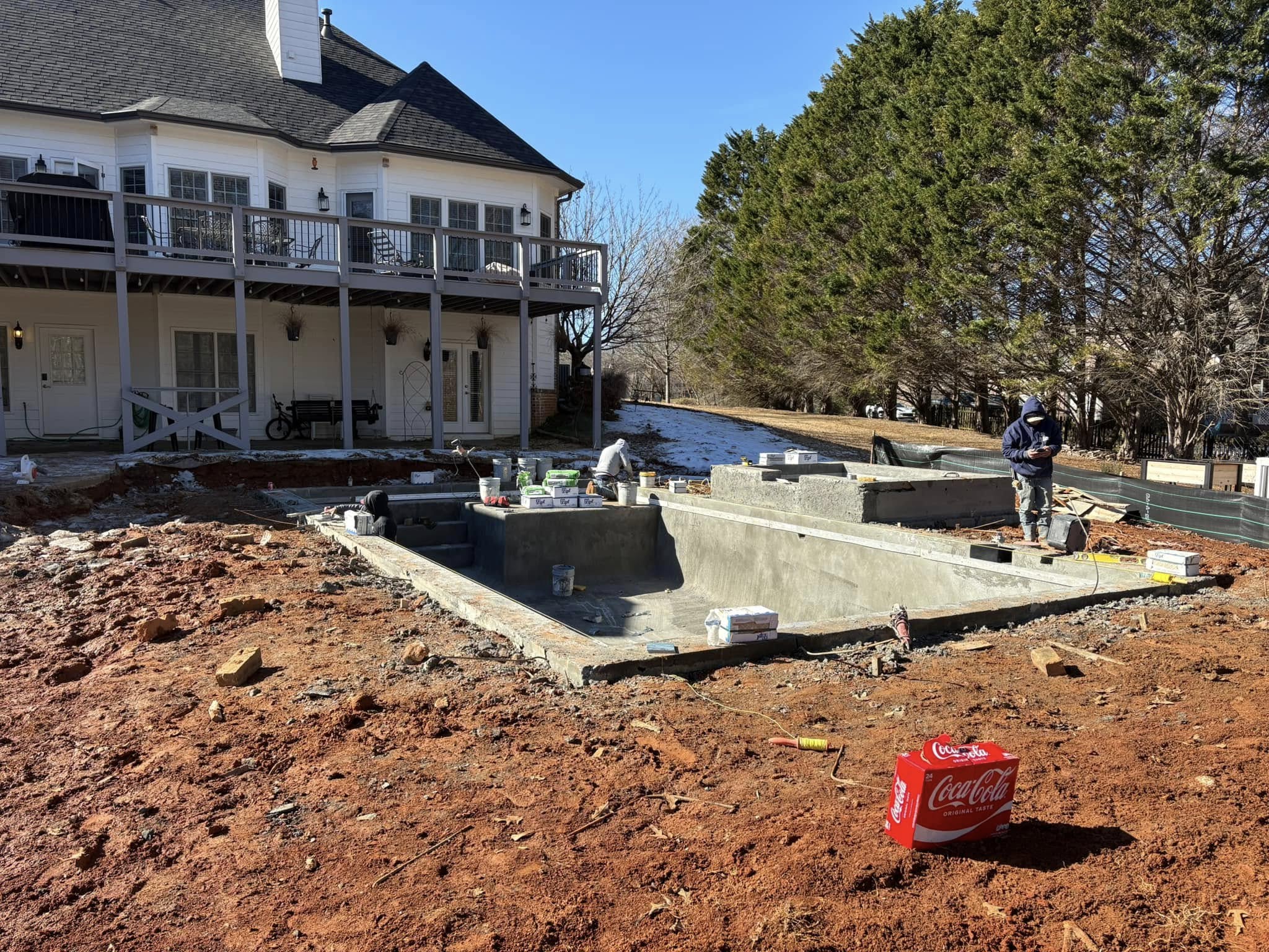 Construction workers build a backyard in-ground pool behind a two-story house, surrounded by red soil and tools. A Coca-Cola box sits in the foreground.