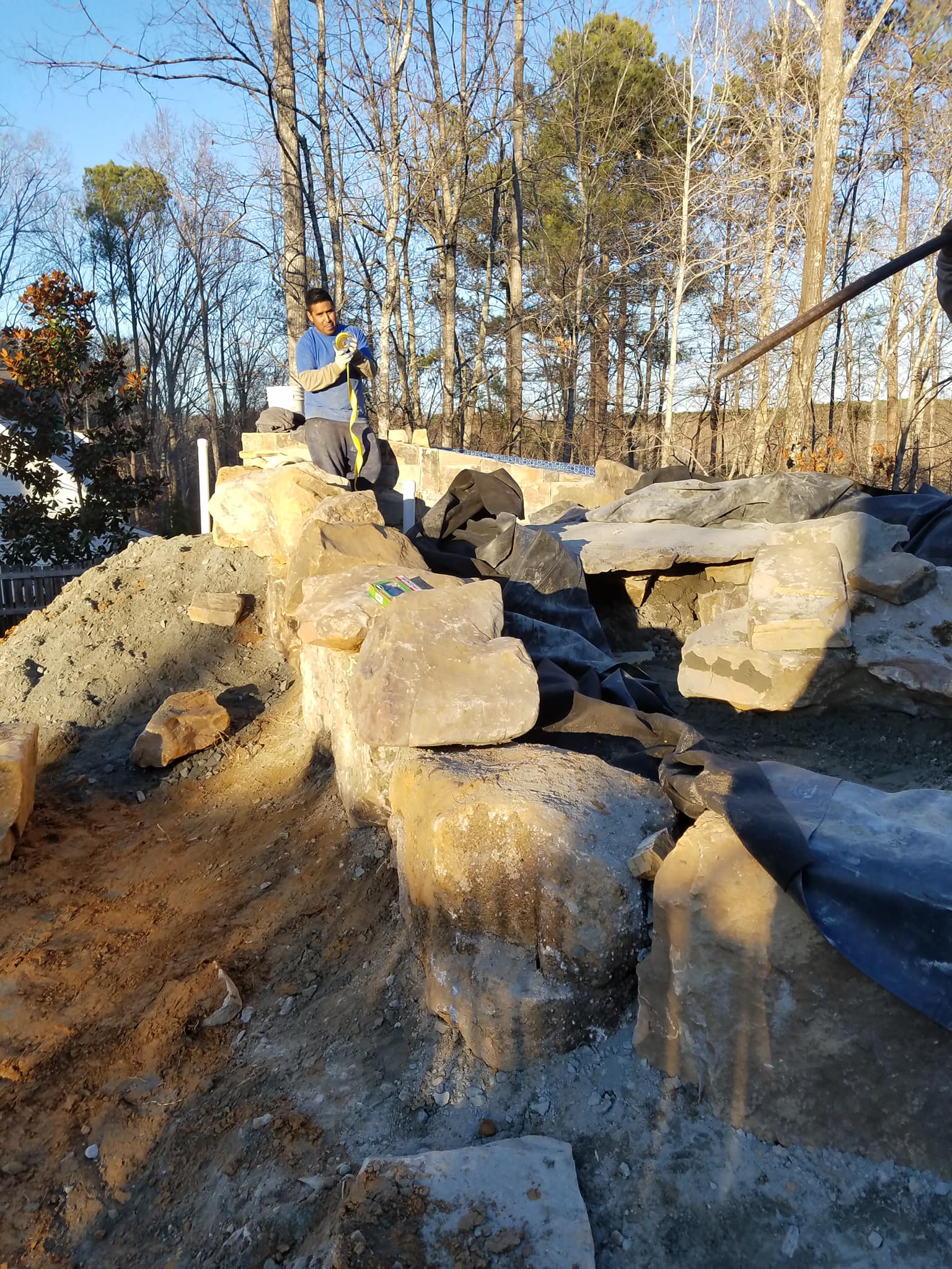 A person working on a rocky outdoor construction site within our service areas, featuring large boulders, a black liner, and surrounding dirt and trees in the background.