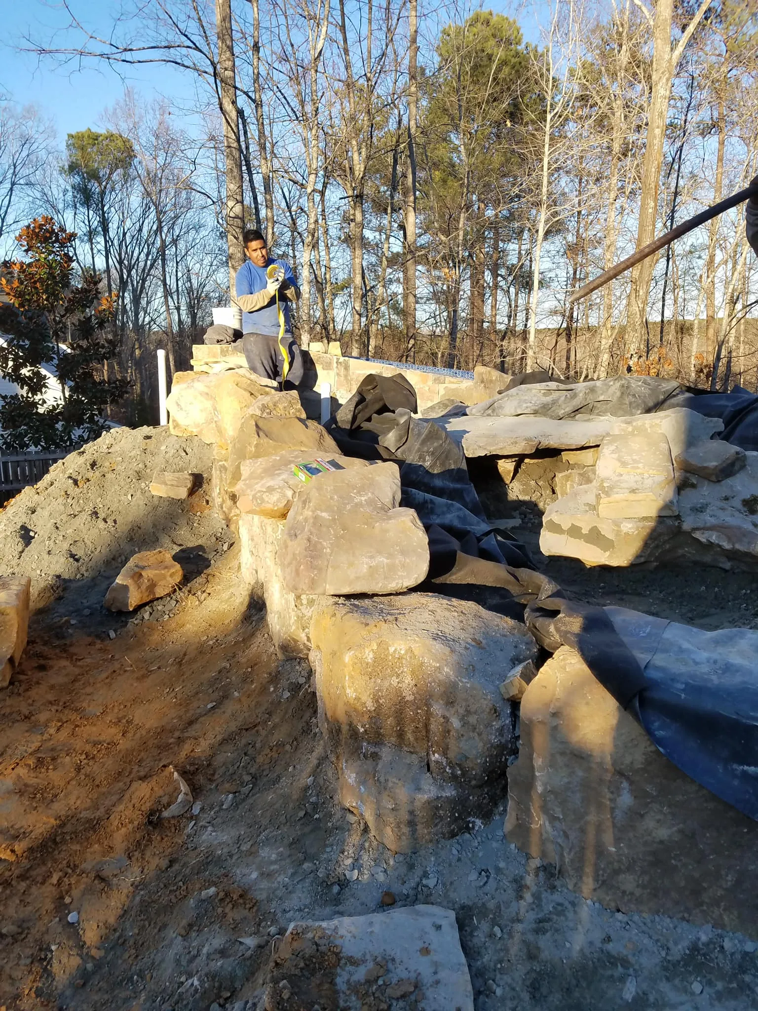 A person working on a rocky outdoor construction site within our service areas, featuring large boulders, a black liner, and surrounding dirt and trees in the background.
