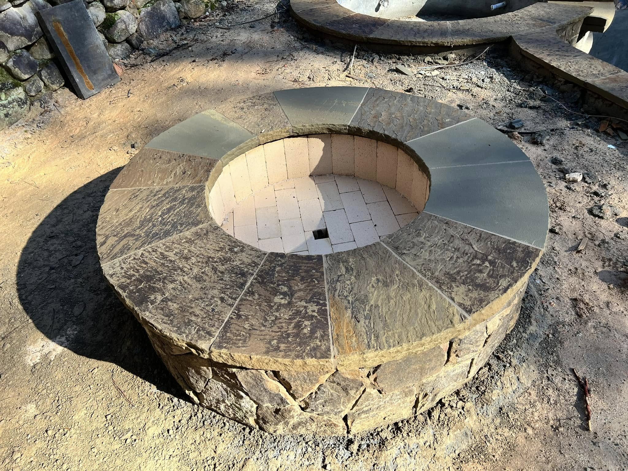 A circular stone fire pit with a brick-lined interior and a small drainage hole at the bottom, set on a dirt ground outdoors.