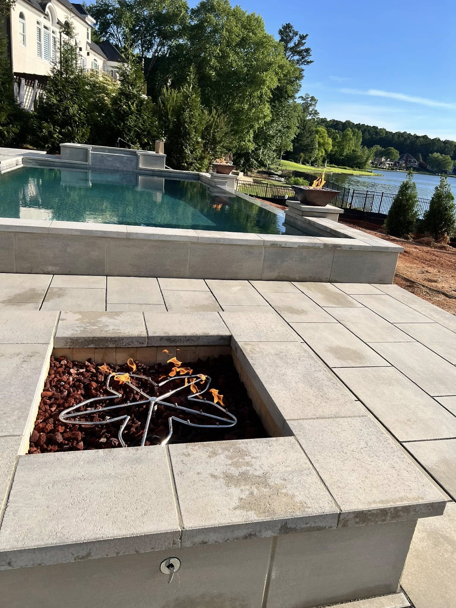 Modern backyard with a rectangular pool, fire pit with visible gas burner, stone patio, and a view of a lake with trees in the background.