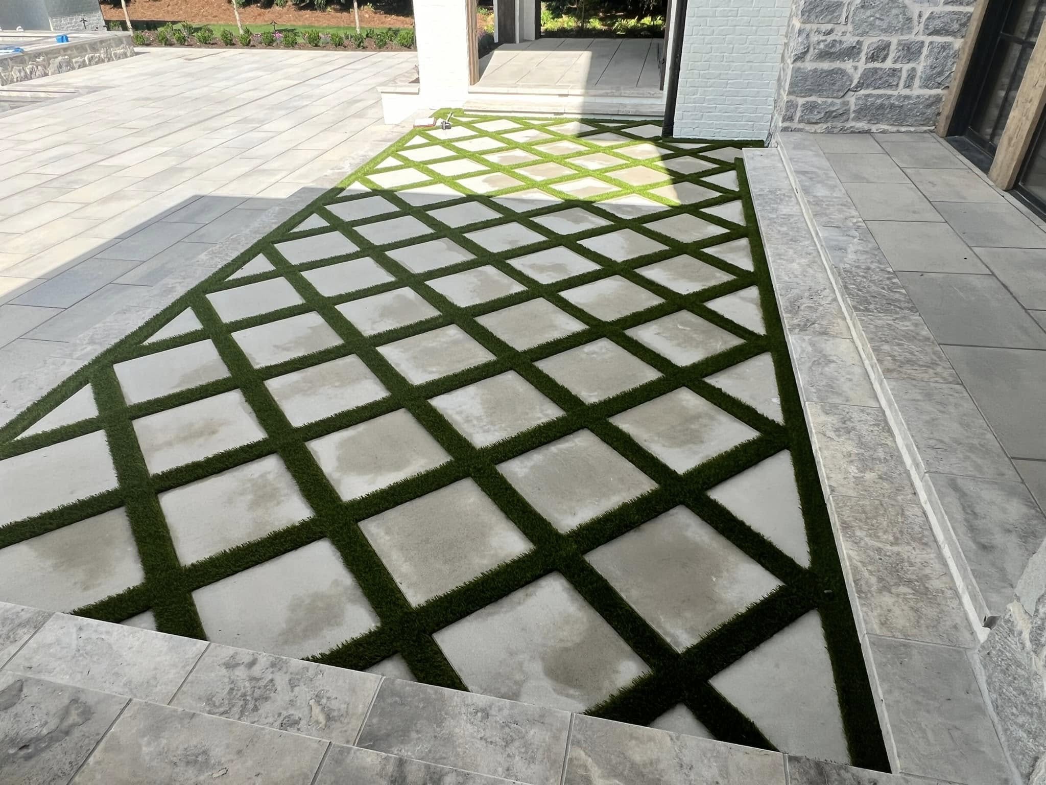 A patio area with square concrete pavers arranged in a diamond pattern, separated by strips of artificial grass, bordered by stone tiles.