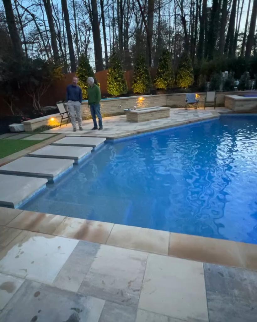 Two people stand by a modern backyard swimming pool with stone steps, surrounded by trees and illuminated by small lights at dusk.
