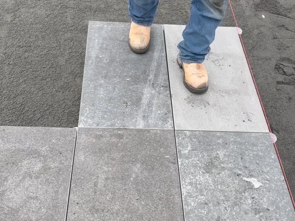 A person wearing tan work boots and blue jeans stands on large gray square tiles being laid on a sand surface.
