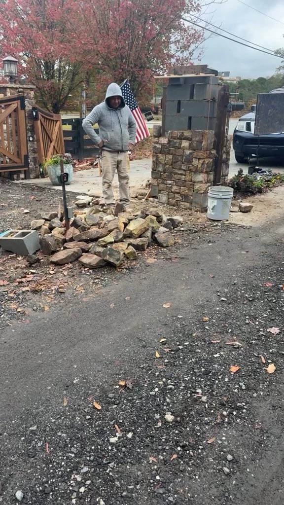 A person in a gray hoodie stands near a pile of rocks and a partially built stone structure beside a road, with an American flag and a parked vehicle in the background.
