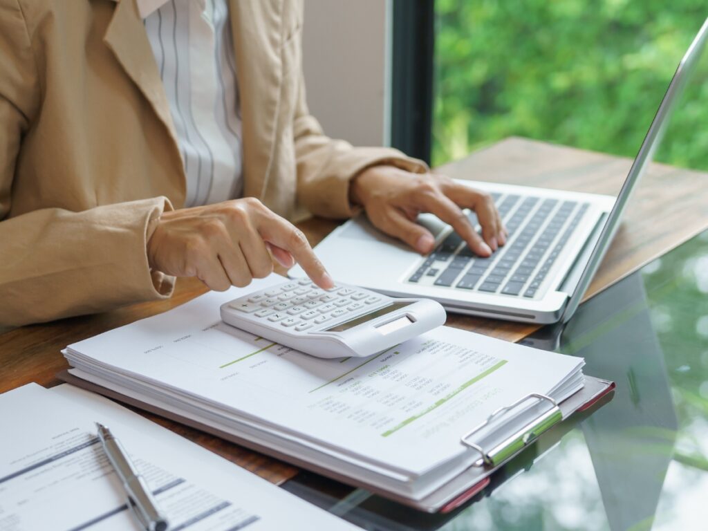 Person in a brown blazer using a calculator and working on a laptop at a desk with financial documents, possibly researching paver patio cost in Georgia, next to a large window with greenery outside.
