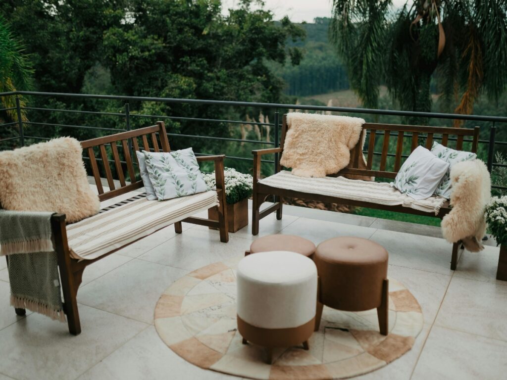 Two wooden benches with cushions and faux fur throws face each other on a tiled patio, complete with round ottomans and a small rug in the center, surrounded by greenery—a stylish setup to inspire your paver patio cost in Georgia planning.