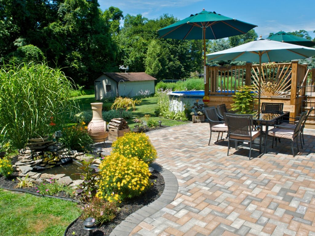 Backyard paver patio with outdoor furniture, large green umbrellas, potted plants, a small pond, and a shed in the background on a sunny day—discover the average paver patio cost in Georgia for spaces like this.