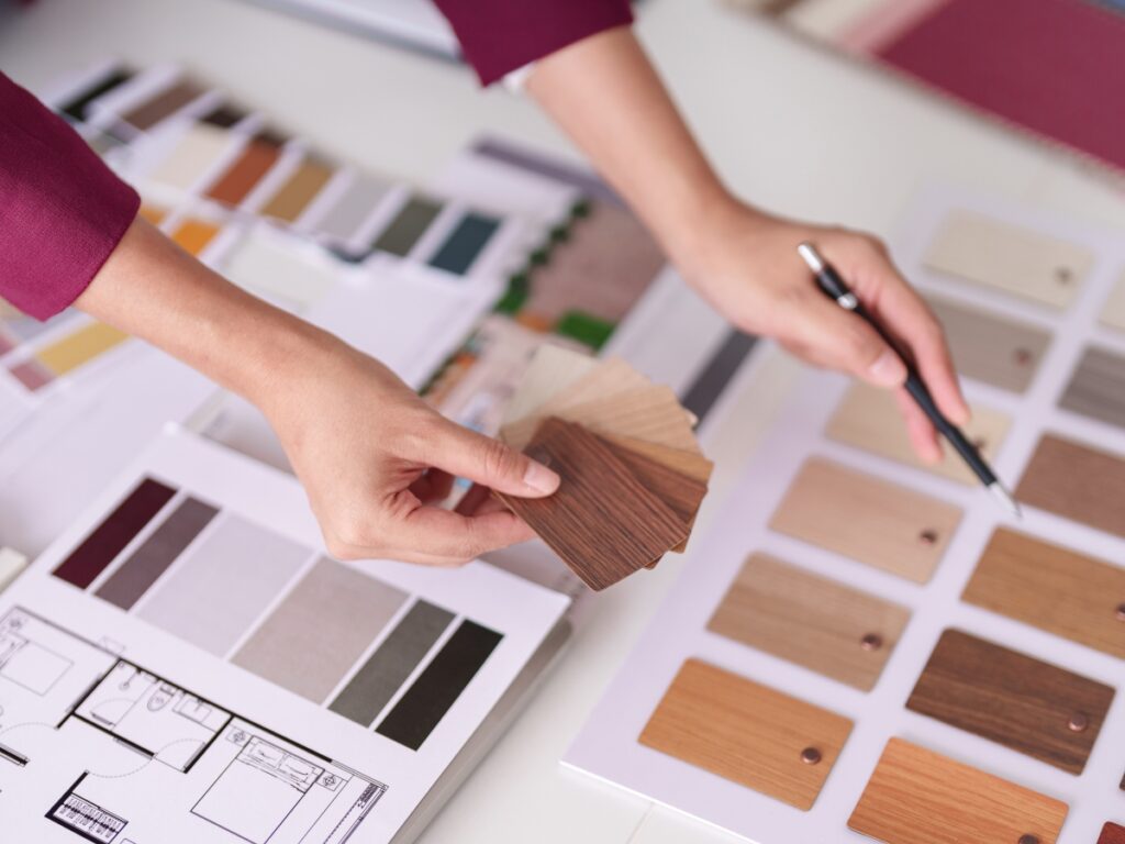 Person comparing wood laminate samples and color swatches on a desk with a floor plan, selecting materials for interior design or renovation while considering options like the paver patio cost in Georgia.
