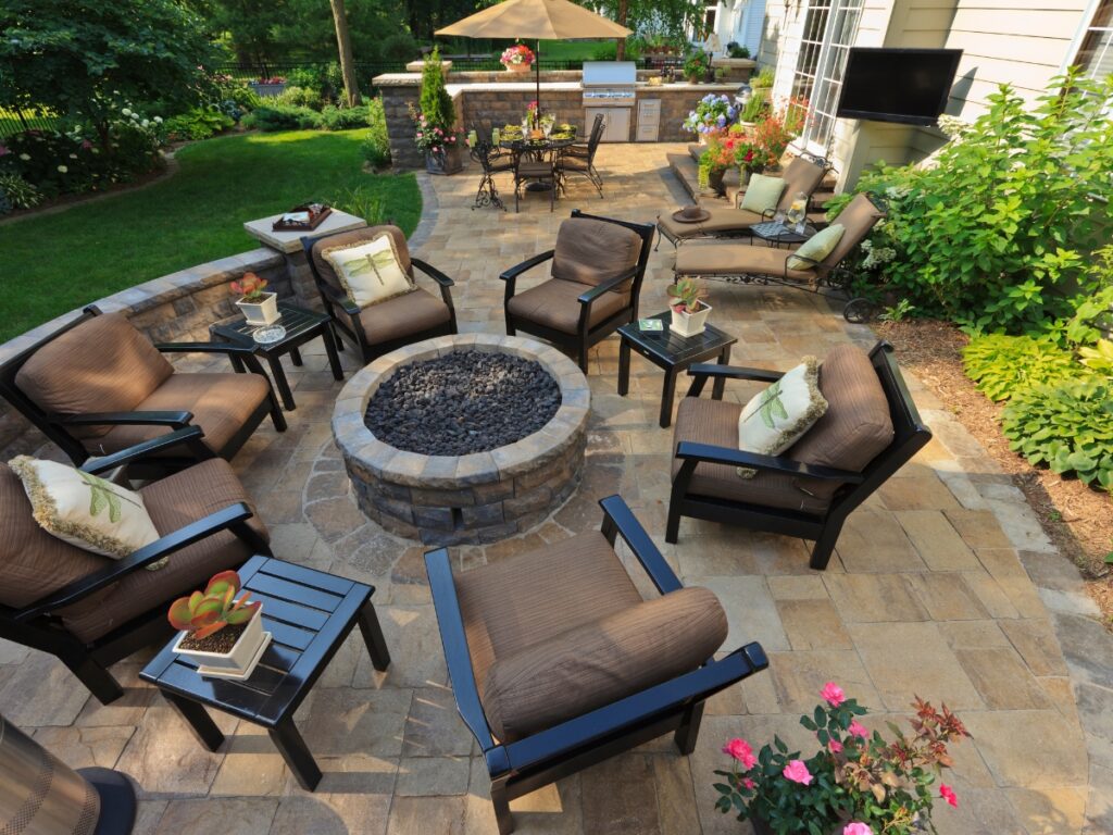 A backyard paver patio with cushioned chairs arranged around a stone fire pit, surrounded by lush plants, small tables, and an outdoor dining area—ideal inspiration when considering paver patio cost in Georgia.